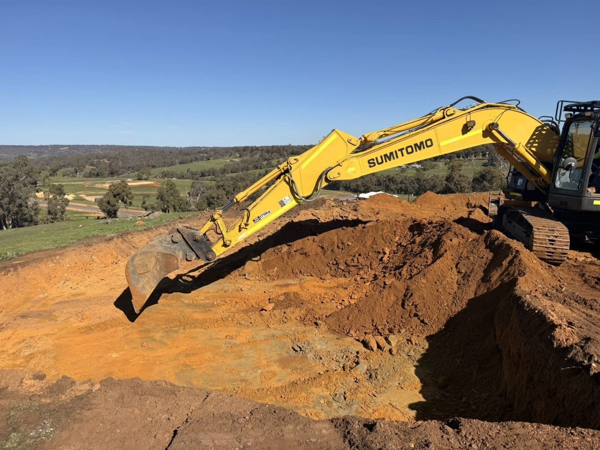 Sumitomo excavator cutting into a red earth batter under a clear blue sky