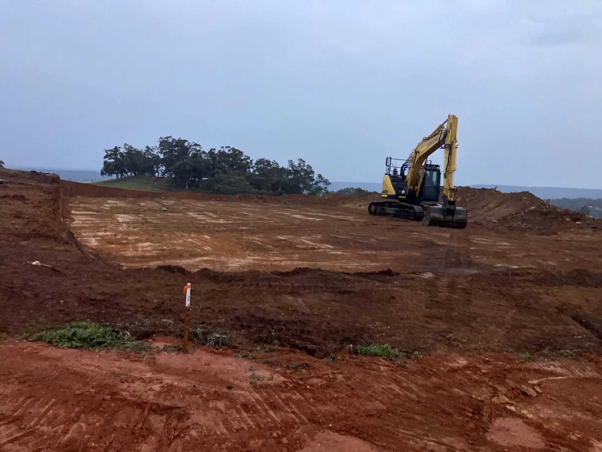 Levelled lot with a survey peg in the foreground and an excavator finishing the pad
