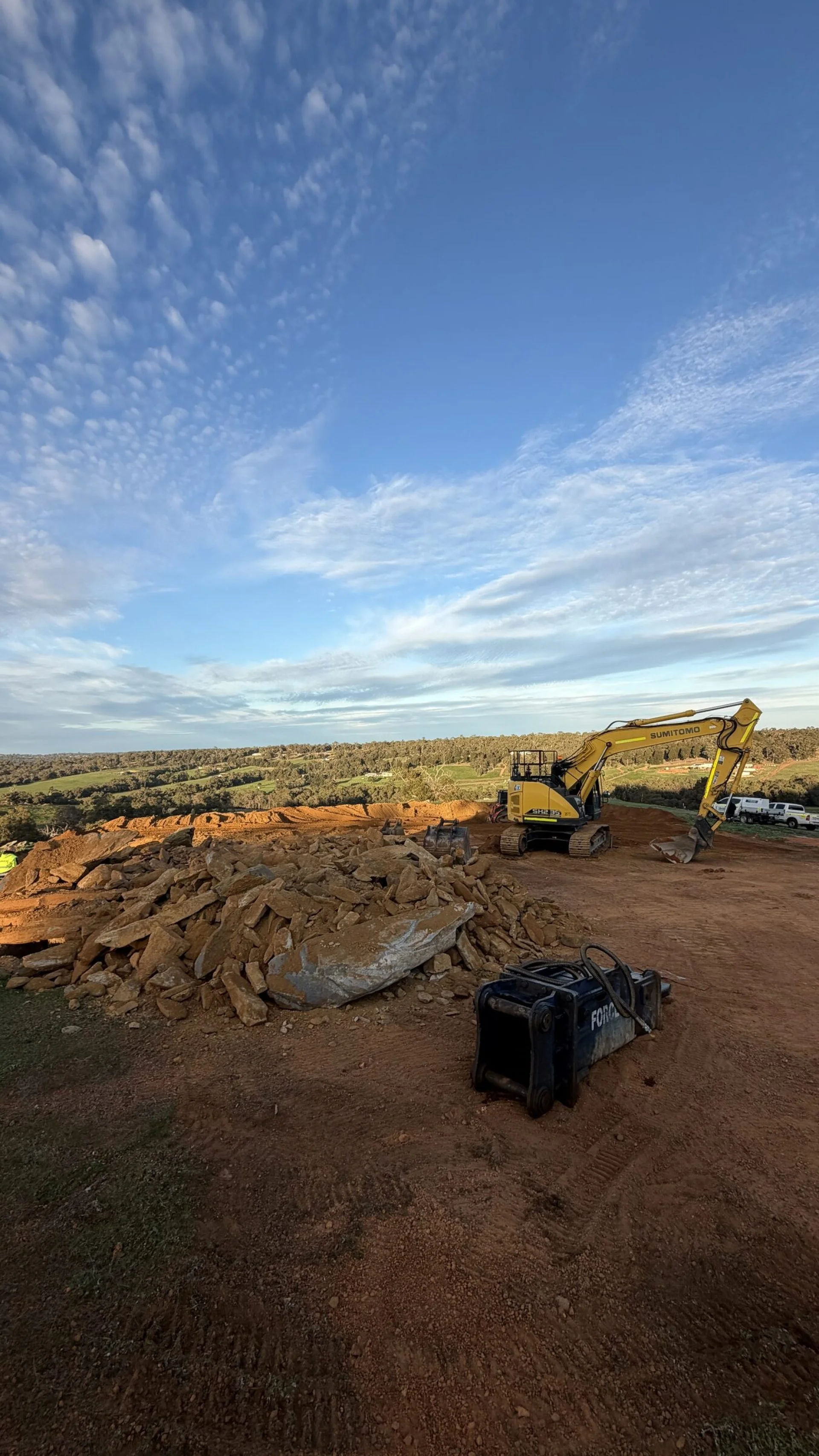 Rock excavation site with a hydraulic breaker in the foreground and an excavator beyond